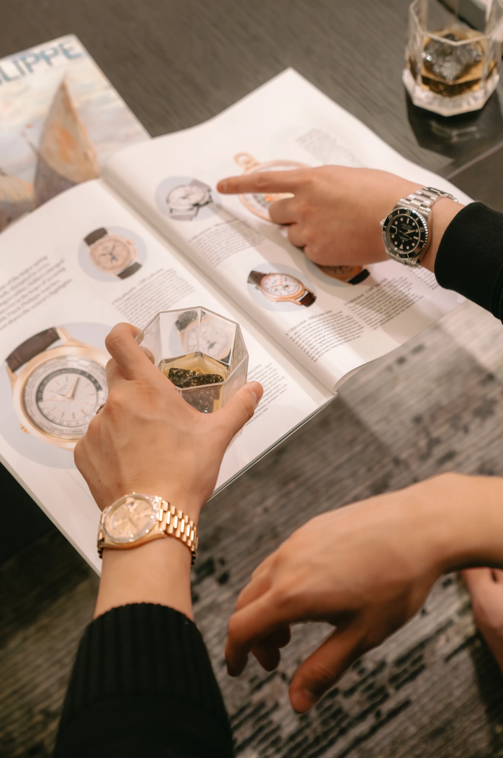 Two collectors talking over a watch reference book, a Submariner and a gold Day-Date on their wrists